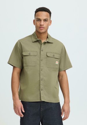 Junger Mann trägt ein olivgrünes, kurzärmliges Hemd mit Knöpfen und zwei Brusttaschen sowie eine dunkle Jeans, steht vor einem schlichten Hintergrund.