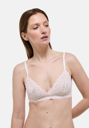 Femme à la peau claire et aux cheveux bruns portant un bralette en dentelle rose pâle, regardant légèrement vers le bas et sur sa gauche, sur un fond uni.
