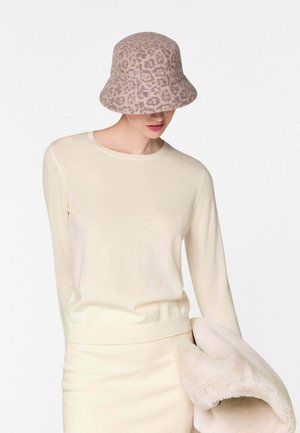 Vrouw draagt een beige top met lange mouwen en een rok, houdt een beige jas van nepbont vast en draagt een roze bucket hat met luipaardprint.