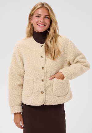 Lächelnde Frau mit langen blonden Haaren trägt eine cremefarbene, strukturierte Jacke mit Knöpfen über einem dunkelbraunen Rollkragenkleid, eine Hand in der Tasche.