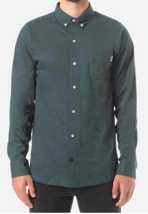 Mężczyzna ubrany w ciemnozieloną, zapinaną na guziki koszulę z długim rękawem i kieszenią na piersi oraz czarne spodnie, stojący na białym tle.