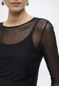 Fekete felső átlátszó, pöttyös mintával, apró gyöngyökkel díszítve. Kerek nyakkivágás, hosszú ujjú, látható pántokkal, amelyek kiemelik a textúrát és a részleteket.
