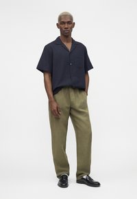 Homme aux cheveux blonds courts portant une chemise à manches courtes bleu marine foncé boutonnée, un pantalon ample vert olive et des chaussures habillées noires, debout sur fond blanc.