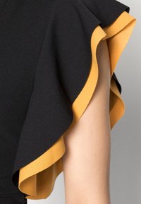Robe noire avec une manche froncée présentant un dessous jaune moutarde contrastant. Texture lisse, forme élégante et bords définis.