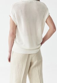 Osoba vnošená volný, krátký rukáv pletený bílý top a béžové široké kalhoty, zobrazena zezadu na bílém pozadí.