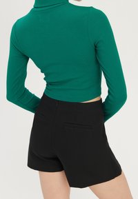 Nainen, joka on pukeutunut vartalonmyötäiseen vihreään ribbipuseroon ja korkeavyötäröisiin mustiin shortsihin, näkyy takaa yksinkertaista taustaa vasten.