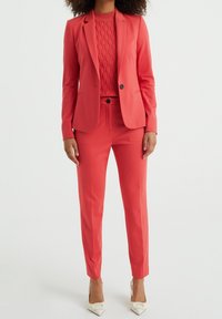 Korallenfarbenes Anzug mit tailliertem Blazer und schmalen Hosen, darunter ein strukturiertes Strickoberteil. Schlichte schwarze Knöpfe und weiße Absatzschuhe.