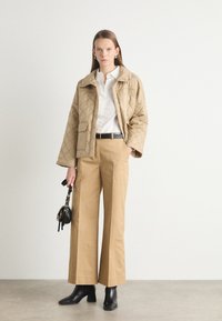 Beige quilted jacket with a pointed collar, paired with cream blouse and wide tan trousers. Black ankle boots and small handbag complete the look.