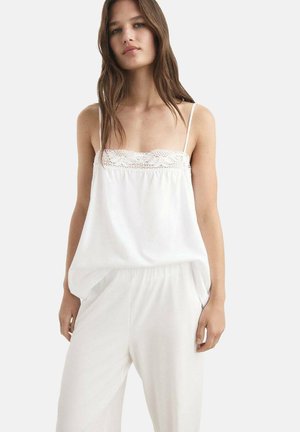 Camisole blanche avec un liseré en dentelle le long de l'encolure, bretelles réglables et une silhouette fluide. Tissu lisse et léger qui tombe librement.
