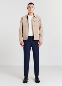 Beige Jacke mit zwei Brusttaschen, weißem T-Shirt, marineblauen Hosen und schwarzen Schuhen. Die Kleidung weist einen modernen Schnitt und eine glatte Textur auf.