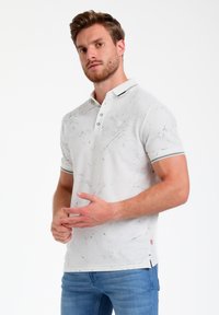 Polo bianco con un motivo testurizzato, maniche corte e colletto a contrasto; abbinato a jeans blu. Presenta bottoni e dettagli laterali.