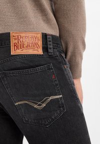 Zwarte denim jeans hebben een bruine leren patch met 'REPLAY BLUE JEANS' branding. De achterzakken zijn afgewerkt met beige stiksel.