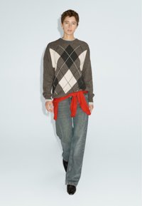 Persona che cammina in avanti indossando un maglione grigio a rombi, una felpa rossa legata in vita, jeans blu e scarpe nere a punta.