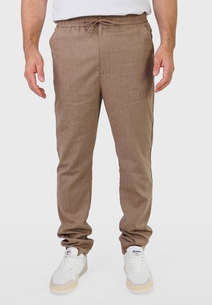 Mand iført lysebrune bukser med snøre og hvide sneakers, der står mod en ensfarvet lys baggrund.