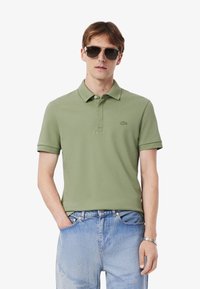 Giovane uomo che indossa una polo verde e occhiali da sole, in piedi con le mani nelle tasche, che mostra uno stile di moda casual.