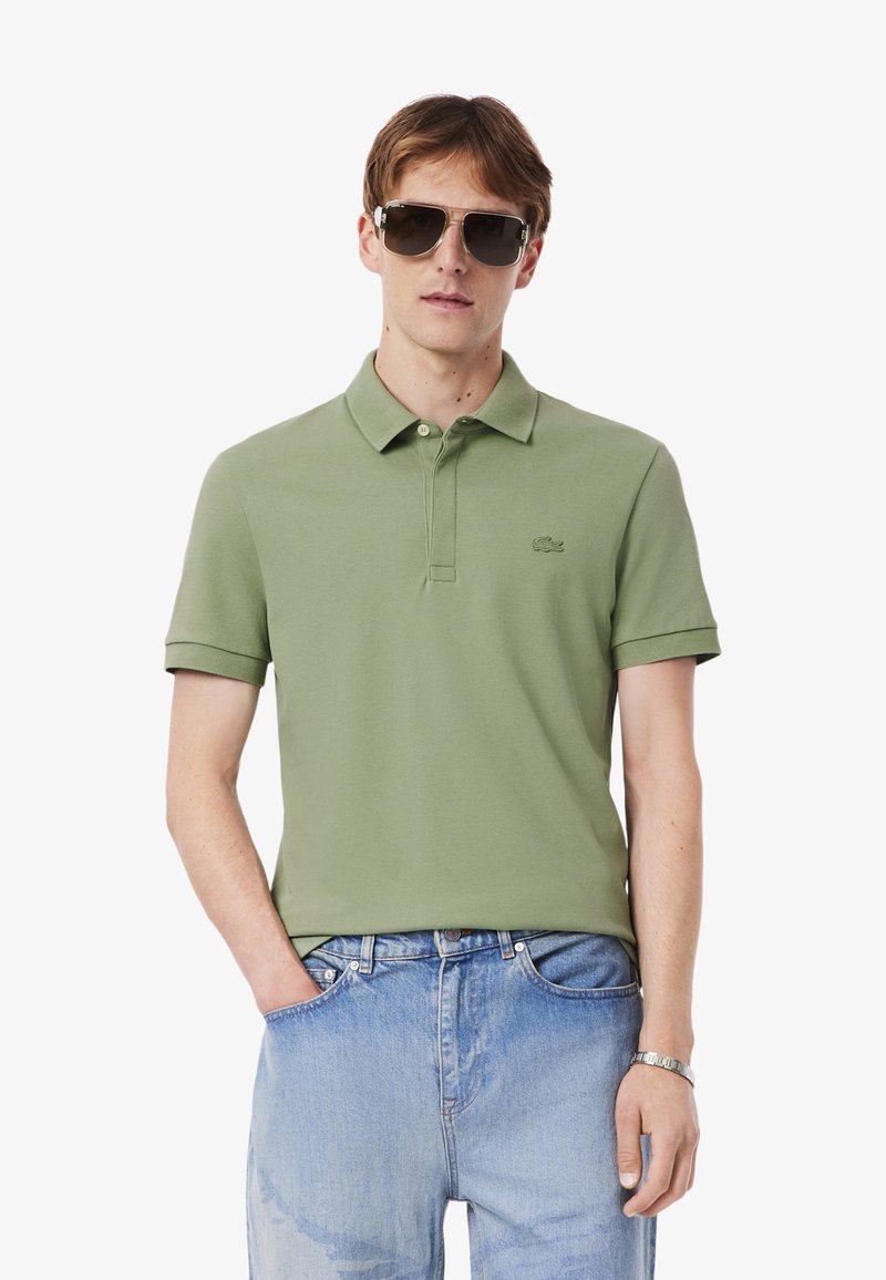 Giovane uomo che indossa una polo verde e occhiali da sole, in piedi con le mani nelle tasche, che mostra uno stile di moda casual.