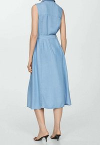 Woman wearing sleeveless light blue denim midi dress with belt, shown from the back, standing in black heeled sandals.