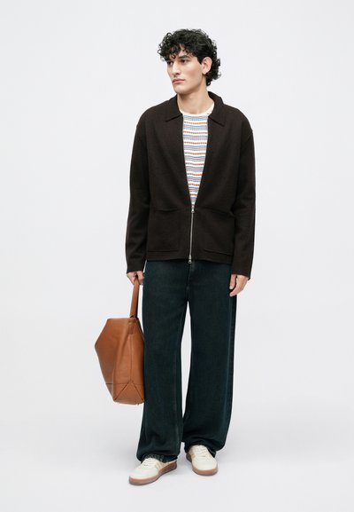 Brązowa kurtka na zamek błyskawiczny na paskowanej koszuli, ciemne jeansy o szerokich nogawkach i białe trampki; trzyma brązową skórzaną torbę typu tote. Jasne tło.