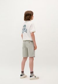 Witte polo met een kleurrijk doodskop- en doodskistenontwerp op de achterkant, gecombineerd met lichtgrijze shorts en witte sneakers.