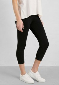 Henkilö, joka pitää yllään mustia lyhyitä legginssejä, valkoisia tenniskenkiä ja löysän vaalean beigepaitaan harmaata seinää ja lattiaa vasten.