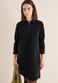 Abito in corduroy nero con zip frontale, colletto a punta, maniche lunghe e due tasche laterali. Texture liscia e design al ginocchio.