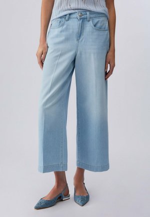 Donna che indossa jeans larghi celeste chiaro e ballerine slingback blu ricamate, in piedi contro uno sfondo neutro.