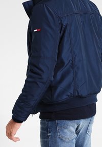 Man som bär en marinblå bomberjacka med en liten rektangulär röd, vit och blå logotyp på den övre vänstra ärmen, tillsammans med ett par blå jeans.