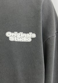 Grå sweatshirts med "Originals Studio" i fed hvid skrift. Stoffet er tekstureret og blødt, med en afslappet pasform og korte ærmer.