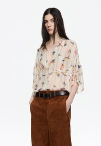 Blusa de gasa beige con patrones florales, con botones en la parte delantera y mangas cortas, combinada con pantalones de ante marrón y un cinturón negro.