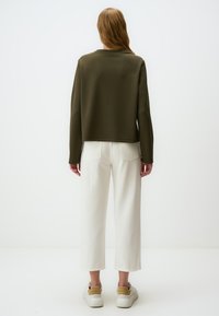 Felpa verde oliva a maniche lunghe e taglio corto, abbinata a jeans bianchi a gamba larga. Il modello indossa sneaker beige e bianche. Design pulito e minimal.