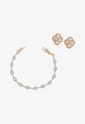 Arany és gyémánt karkötő ovális láncszemekkel, arany és gyémánt virág alakú pötty fülbevalóval egy fehér háttéren.