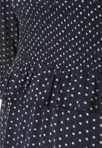 Marinblått tyg med knäppt byst har vita, oregelbundna prickar. Designen inkluderar en volangsläpp och strukturerad rynkning genom hela plagget.