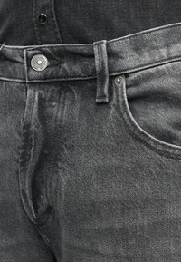 Gros plan sur un jean en denim noir montrant la taille, la boucle de ceinture, la poche avant et un bouton métallique marqué "DONDUP."