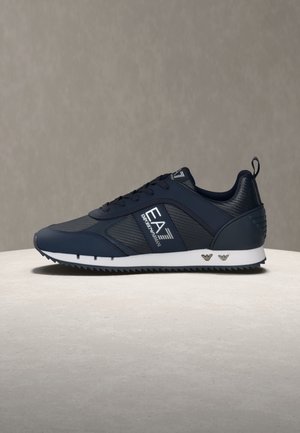 Tumši zils EA7 Emporio Armani sporta apavu pāris ar melnu sietiņu, baltu zolīti, priekšpusē ar šņorēm un logotipu sānos un papēdī, uz neitrālas virsmas.