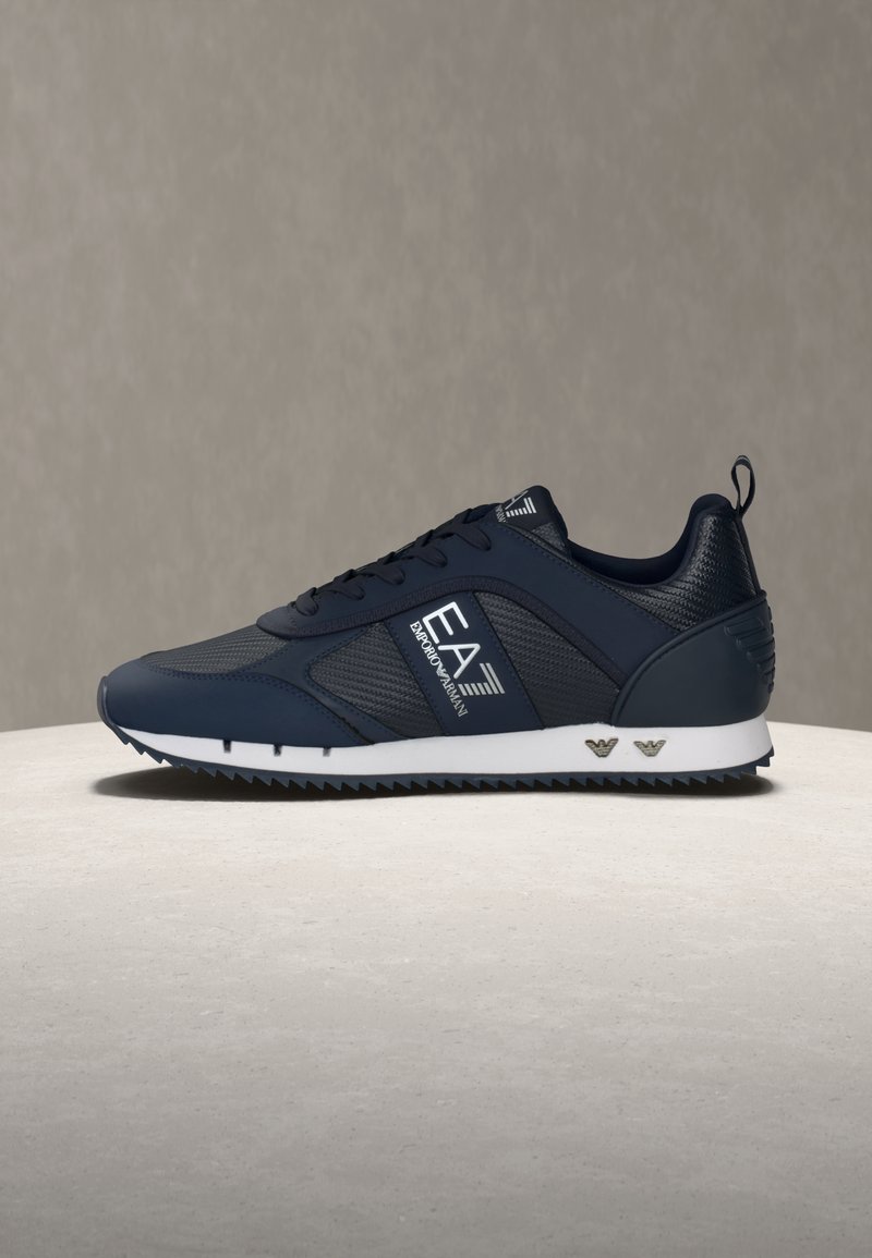 Σκούρο μπλε αθλητικό παπούτσι EA7 Emporio Armani με μαύρο δίχτυ, λευκή σόλα, μπροστινό δέσιμο με κορδόνια και λογότυπο στο πλάι και στη φτέρνα, σε ουδέτερη επιφάνεια.