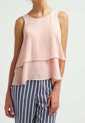 Woman wearing a sleeveless layered light pink blouse and blue pants with vertical white stripes, standing against a plain background.