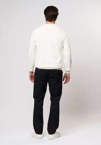Chemise blanche à manches longues, pantalon noir et chaussures blanches. La chemise a un col ras du cou, et le pantalon possède des poches latérales avec des accents en métal doré.