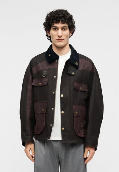 Άνδρας που φορά σκούρο καφέ μπουφάν με πολλαπλές τσέπες με κουμπιά, μαύρο γιακά, λευκό πουκάμισο και γκρι παντελόνι, στέκεται μπροστά σε απλό φόντο.