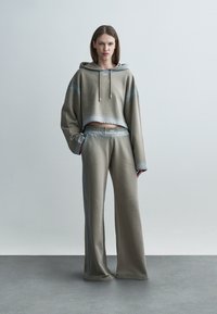 Γυναίκα που στέκεται σε γκρι τοίχο, φοράει γκρι φαρδιά κοντή φούτερ μπλούζα με κουκούλα και παντελόνι με φαρδιά πόδια, με λογότυπα "OFF" στο στήθος και τη μέση.