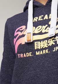 Sudadera con capucha en tejido gris oscuro con cordones blancos. Presenta el colorido logo "Superdry" en degradado de rosa a amarillo, con detalle de etiqueta.