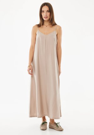 Mujer con un vestido largo sin mangas suelto de color beige claro con tirantes finos y zapatos beige perforados con cordones verdes, de pie sobre un fondo blanco.