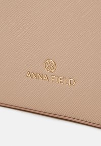 Sac à main beige texturé avec un logo "ANNA FIELD" en relief doré et un motif géométrique subtil sur la surface.