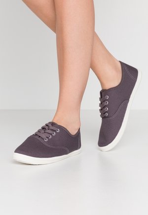 Donkeregrijze canvas sneakers met witte rubberen zolen, vetersluiting, ronde neus en subtiele stikdetails. Voet gedeeltelijk zichtbaar.