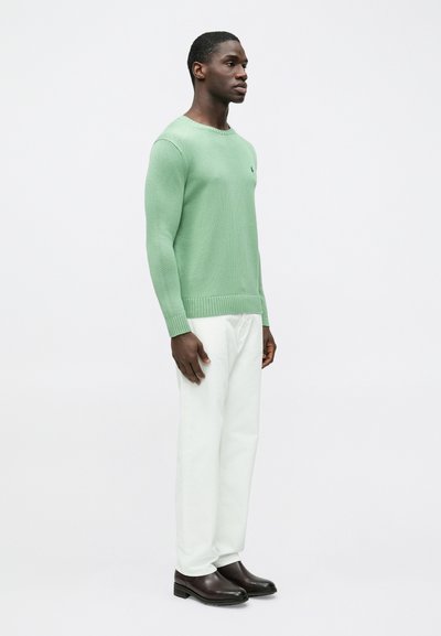 Mint zelena pletena majica z rebrastim spodnjim robom, kombinirana z belimi hlačami in temno rjavimi škornji. Model stoji v profilu na belem ozadju.