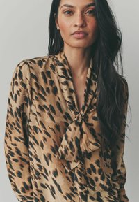 Blouse en soie imprimée léopard beige clair avec un détail de nœud au cou, ornée de taches noires et d'une texture lisse. Manches longues, coupe décontractée.