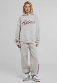 Svijetlosiva oversized dukserica s ružičastim vezom "Felicious" logotipom, u kombinaciji s trenirkama koje imaju butterfly ukrase i manžete na gležnjevima.