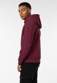 Burgunderfarbener Hoodie mit Kängurutasche, verstellbarem Kapuzenzug und gerippten Bündchen. Schwarzes Logo auf der Rückseite, kombiniert mit schwarzen Cargo-Hosen.