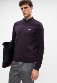 Lila Strickpullover mit geripptem Design, Rundhalsausschnitt und BOSS-Logo. Das Model hält eine schwarze Tasche und trägt graue Hosen.