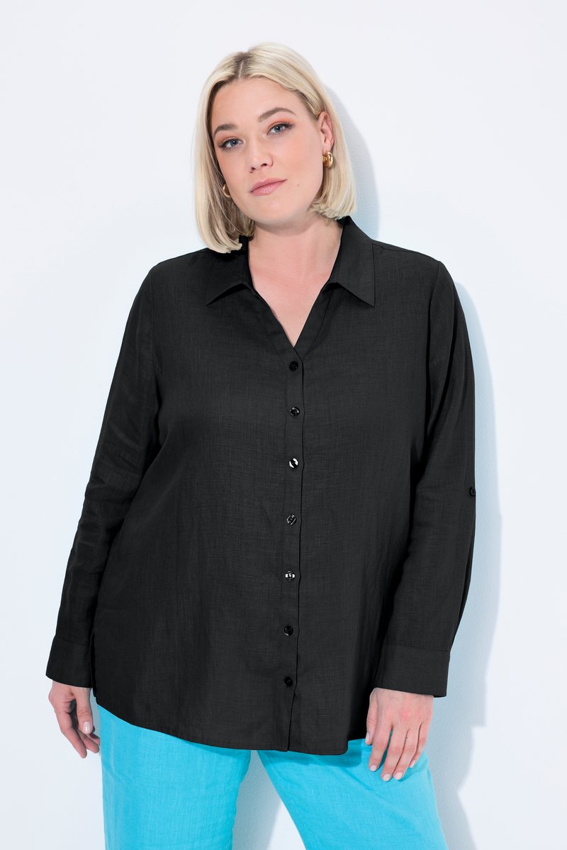 Femme aux cheveux blonds courts portant une chemise noire boutonnée et un pantalon bleu vif, debout sur un fond blanc.