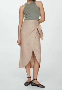 Vrouw in een mouwloze grijze top, beige wikkelrok aan de zijkant geknoopt, en zwarte mesh flats met enkelslierten, staand tegen een effen achtergrond.